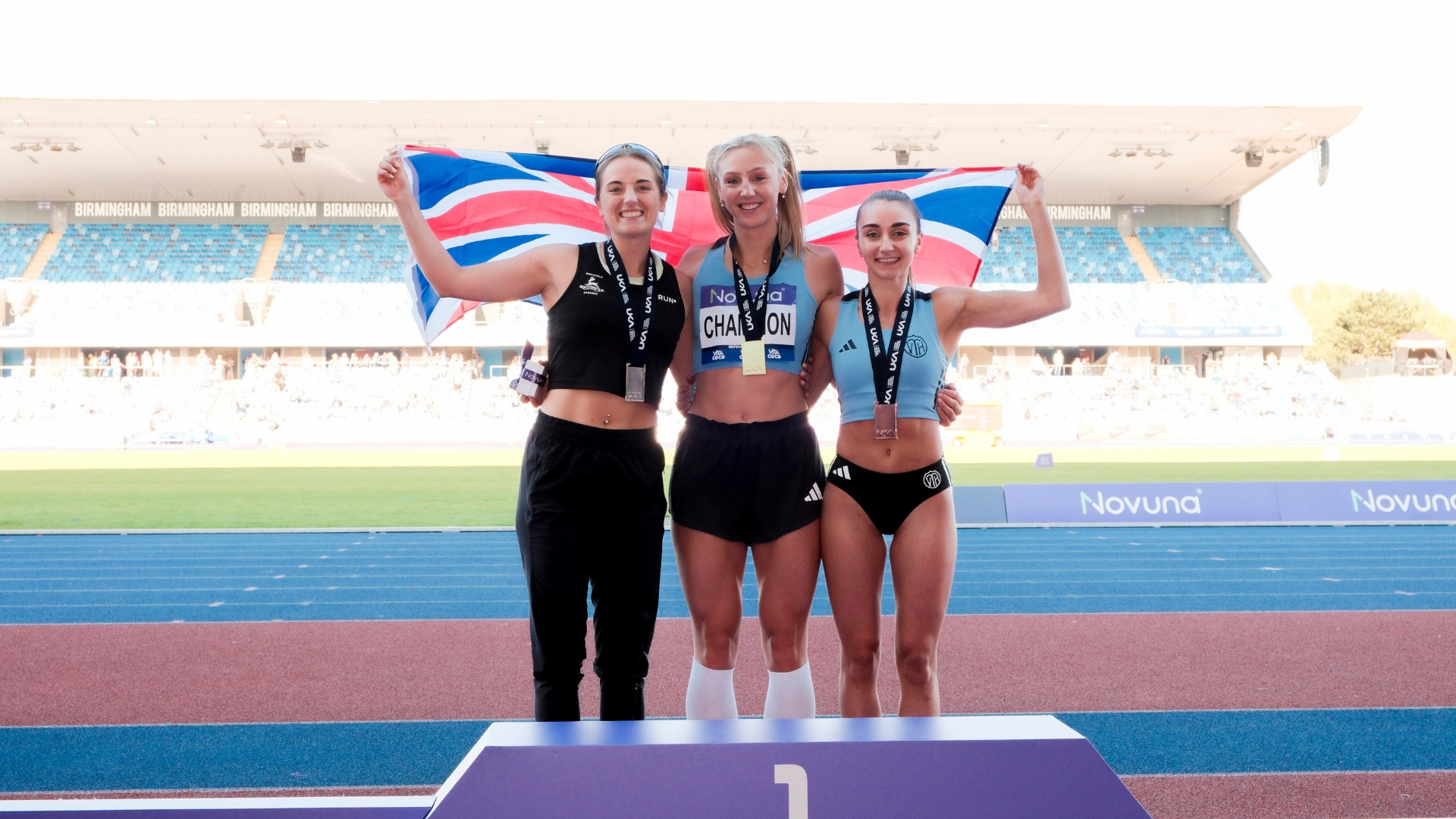 Three medal-winning athletes standing on the podium at a track championship, smiling and holding a Union Jack flag behind them in the stadium.