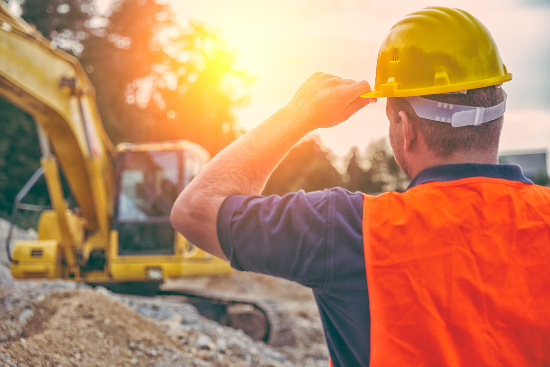 Construction worker wearing a hard hat and orange safety vest overlooking a dig site with a yellow excavator in bright afternoon sun.