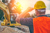 Construction worker wearing a hard hat and orange safety vest overlooking a dig site with a yellow excavator in bright afternoon sun.
