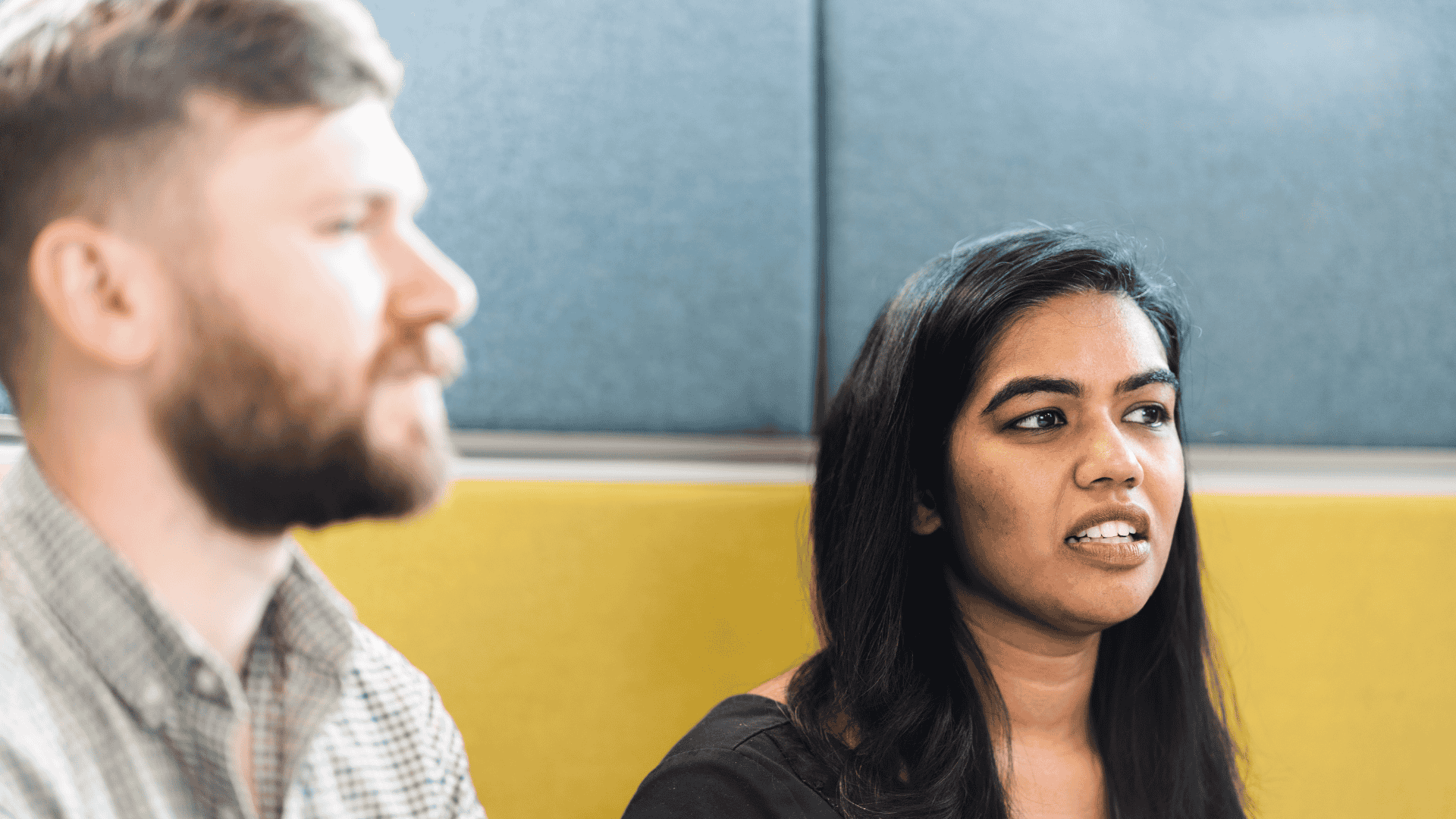 Two colleagues in discussion, with a woman speaking in the foreground and a man listening beside her in a modern office setting.