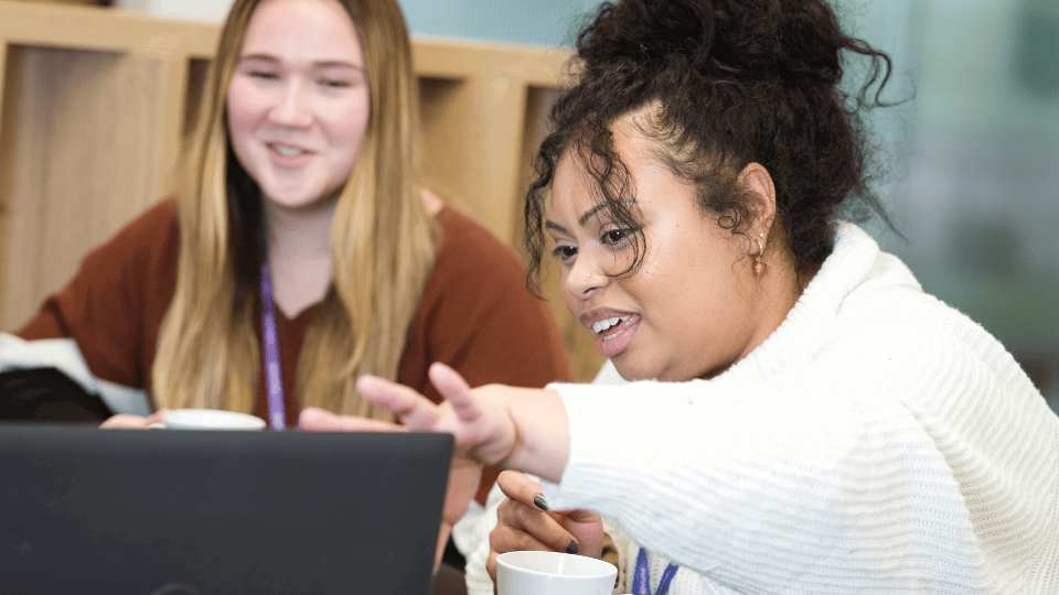 Two colleagues collaborating at a laptop, with one woman pointing at the screen while the other smiles beside her