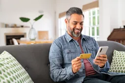 Man sitting on a sofa with green cushions buying something on his phone with a credit card