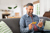 Man sitting on a sofa with green cushions buying something on his phone with a credit card
