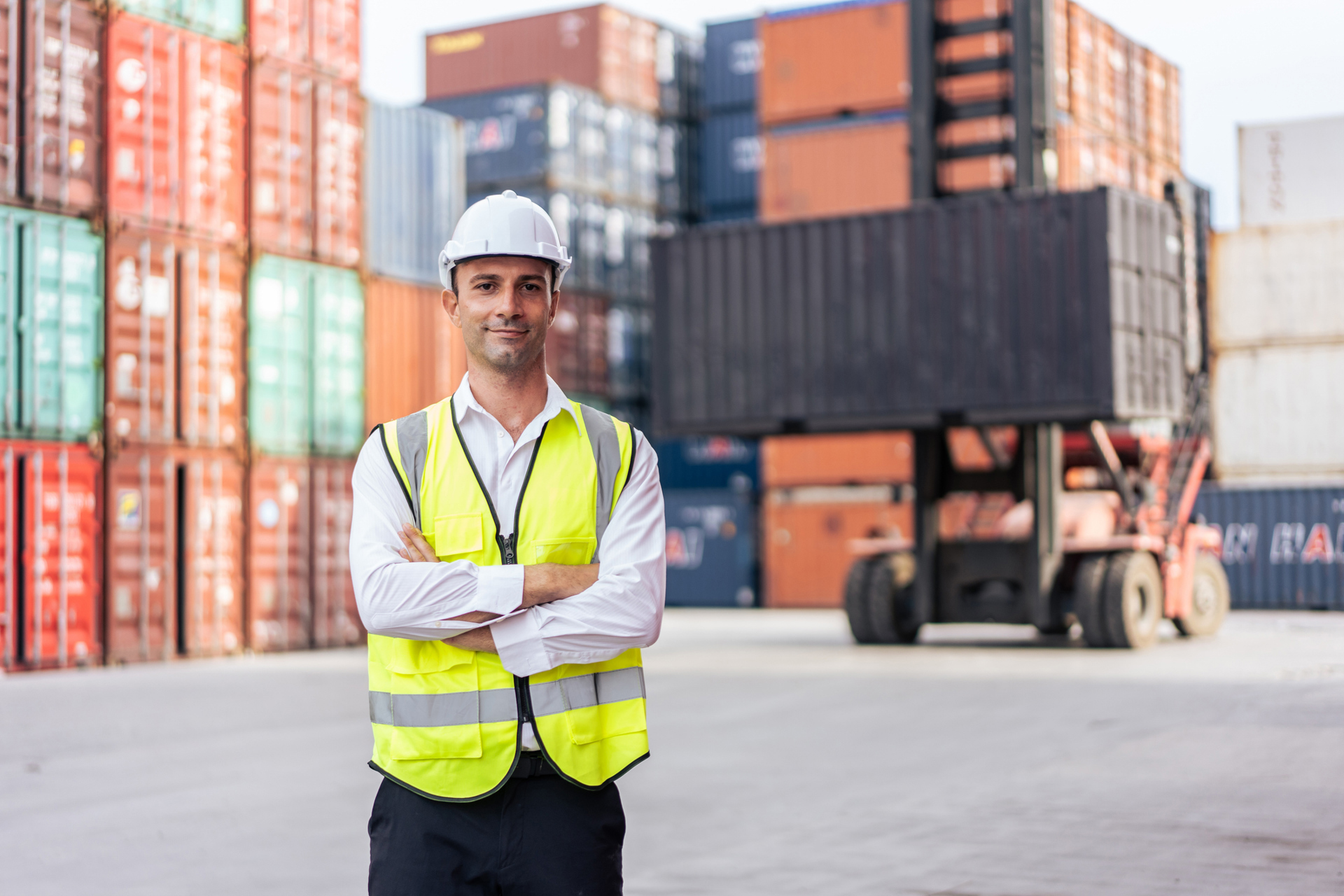 Logistics supervisor wearing a hard hat and hi-vis vest standing confidently in front of stacked shipping containers and a container forklift at a busy freight yard