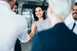 Businesswoman shaking hands with a man in a white shirt, with colleagues standing around them