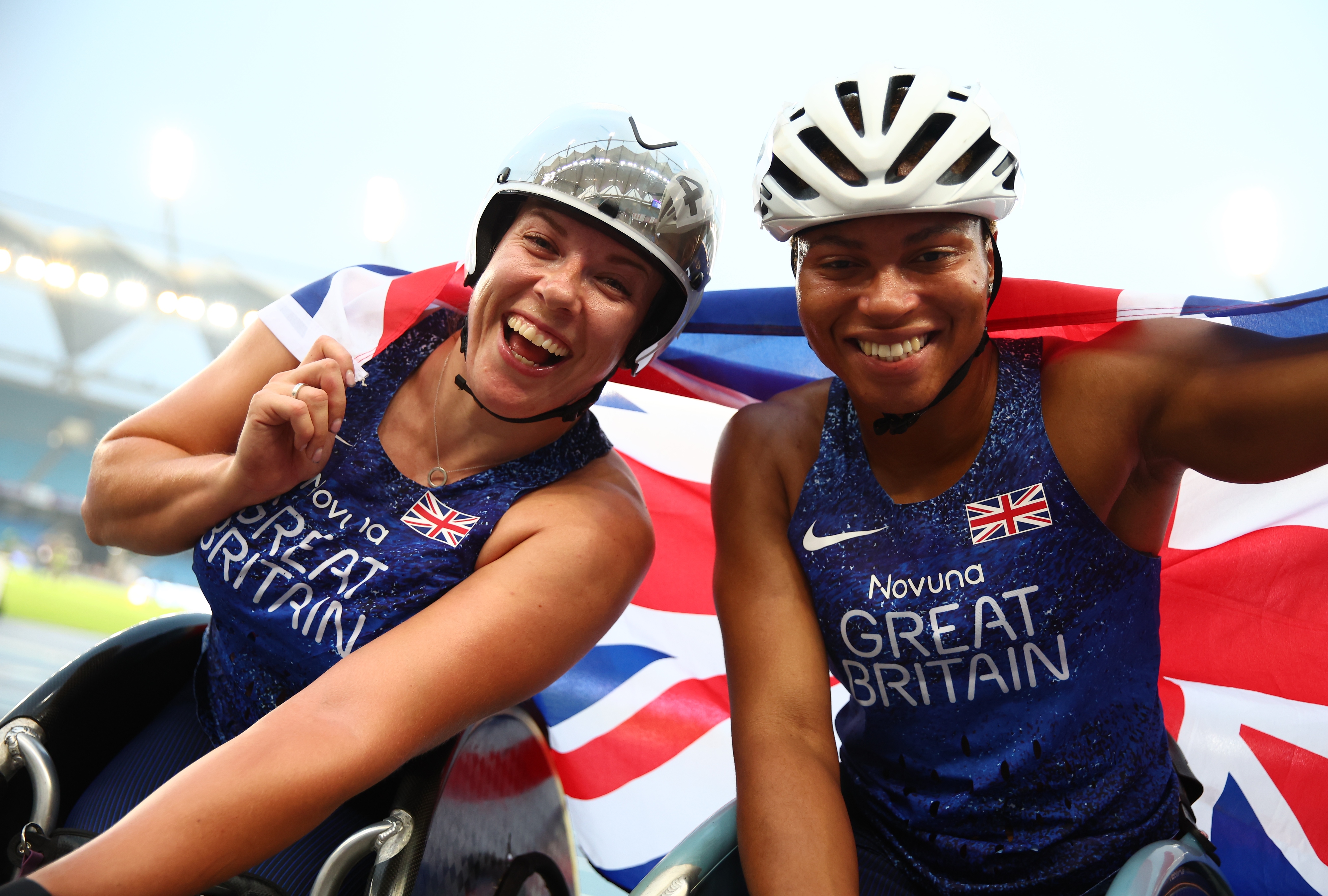 Two Great Britain wheelchair racers smiling and celebrating on the track, holding a Union Jack flag behind them after a race.
