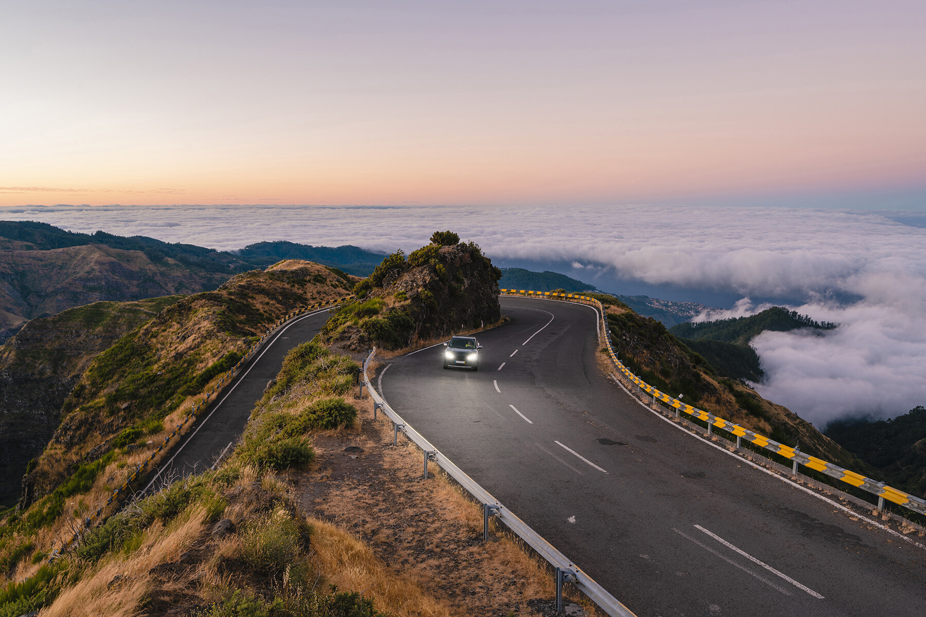 Car driving along a winding mountain road at sunrise, surrounded by dramatic peaks and low-lying clouds