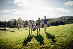 Three men walking across a golf course in the sunshine carrying their equipment
