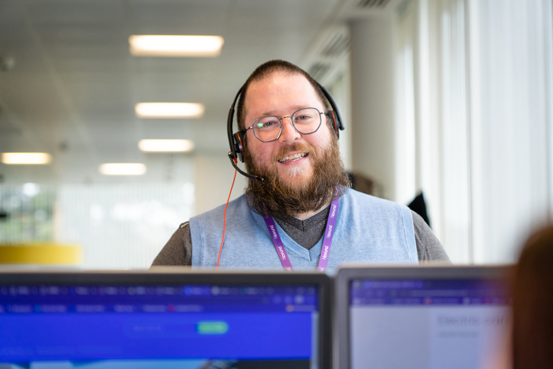 Smiling customer service team member wearing a headset and Novuna lanyard, working at a computer in a bright office